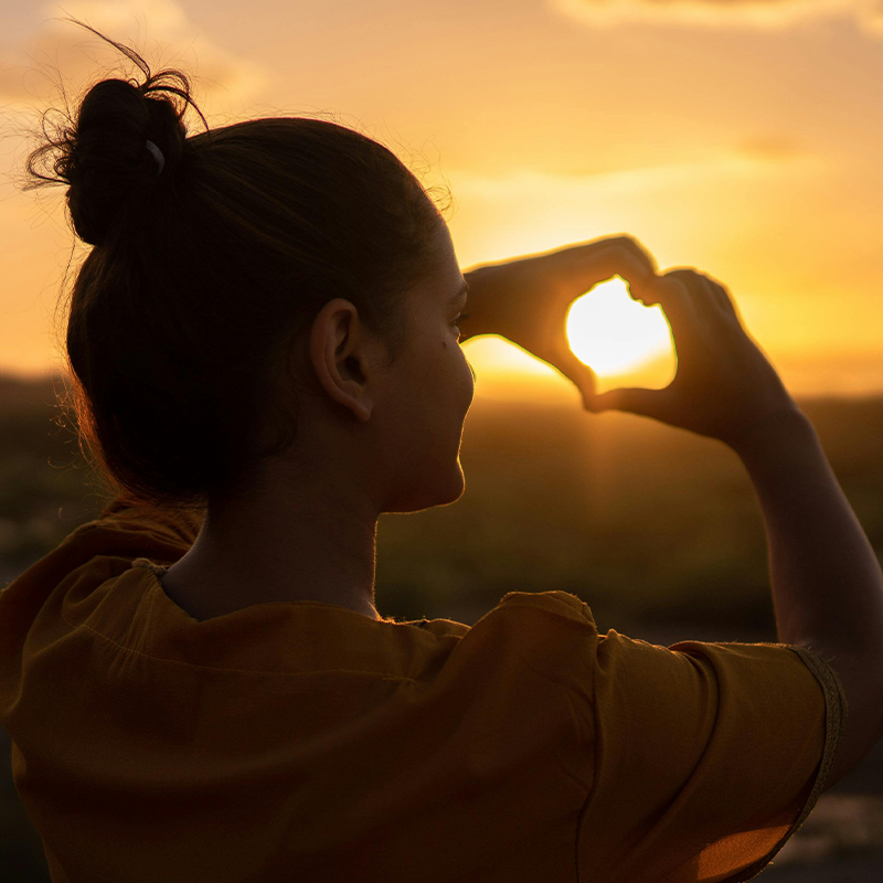 Stress Management, San Diego, Image of a happy woman holding heart hands at the sun.